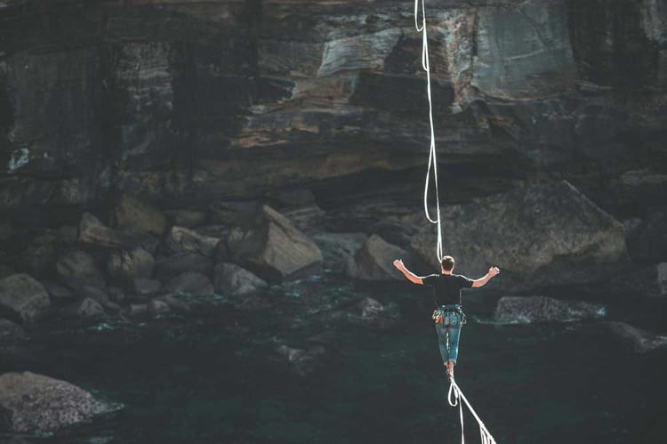 A person walking on a tightrope suspended over a rocky area near the water.