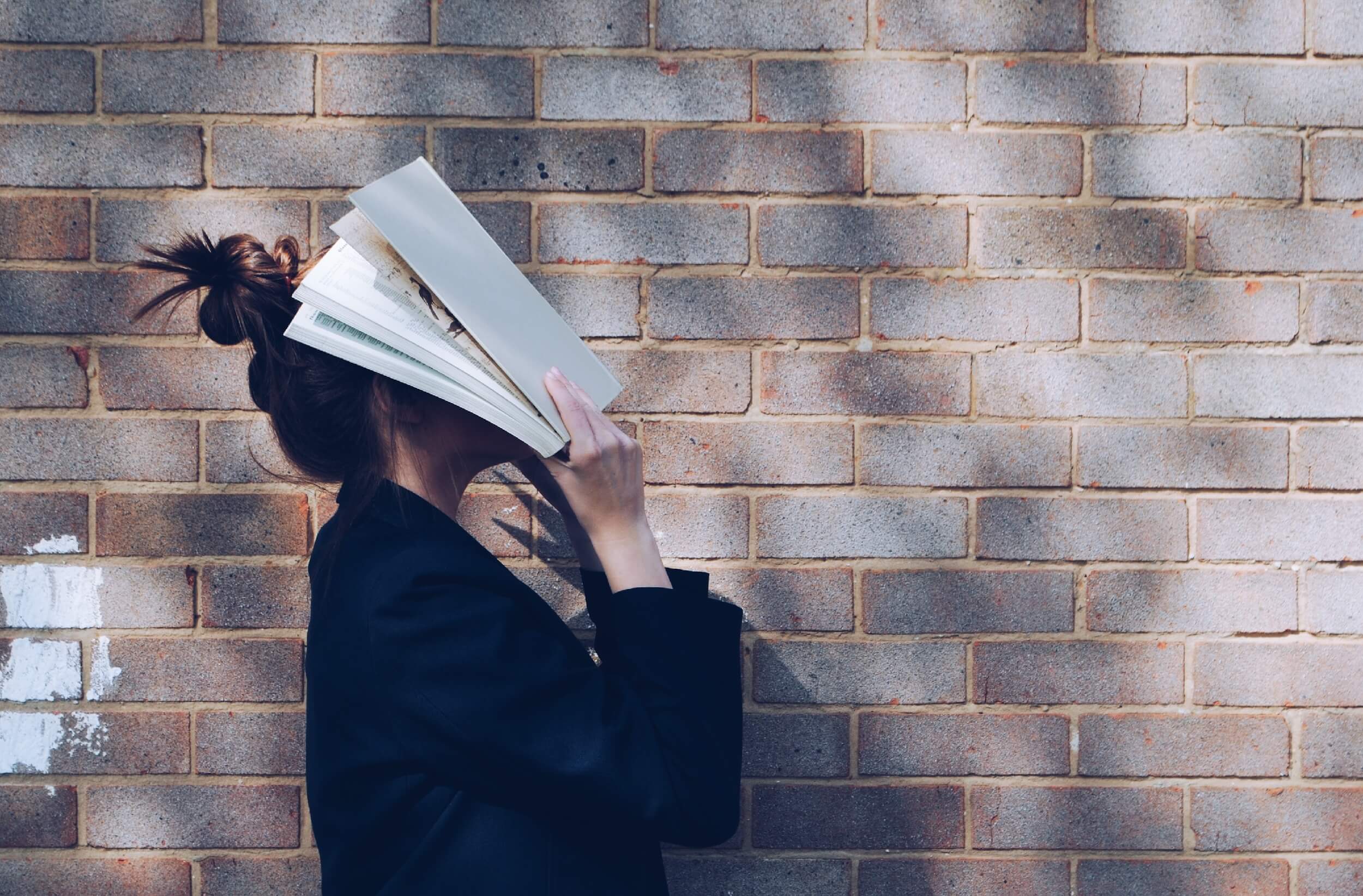 A person standing against a brick wall, holding a stack of books in front of their face.